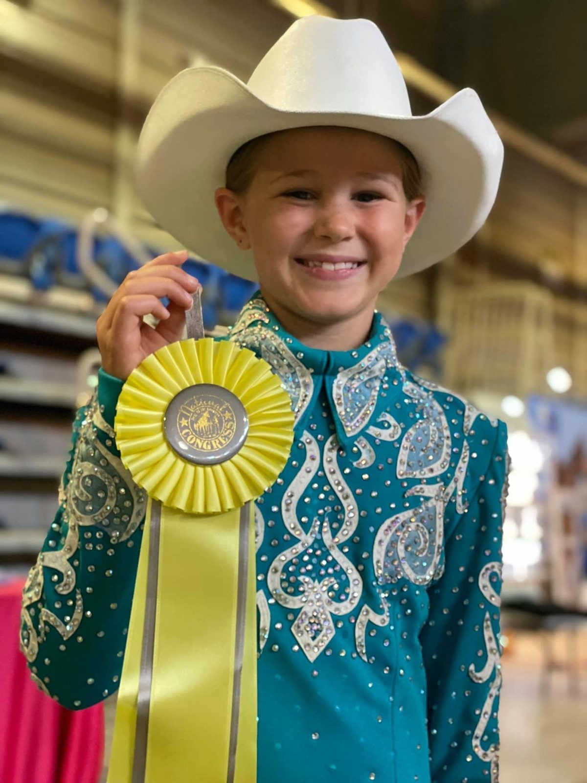 A smiling child in a white cowboy hat and sparkly show shirt holds up a rosette ribbon.