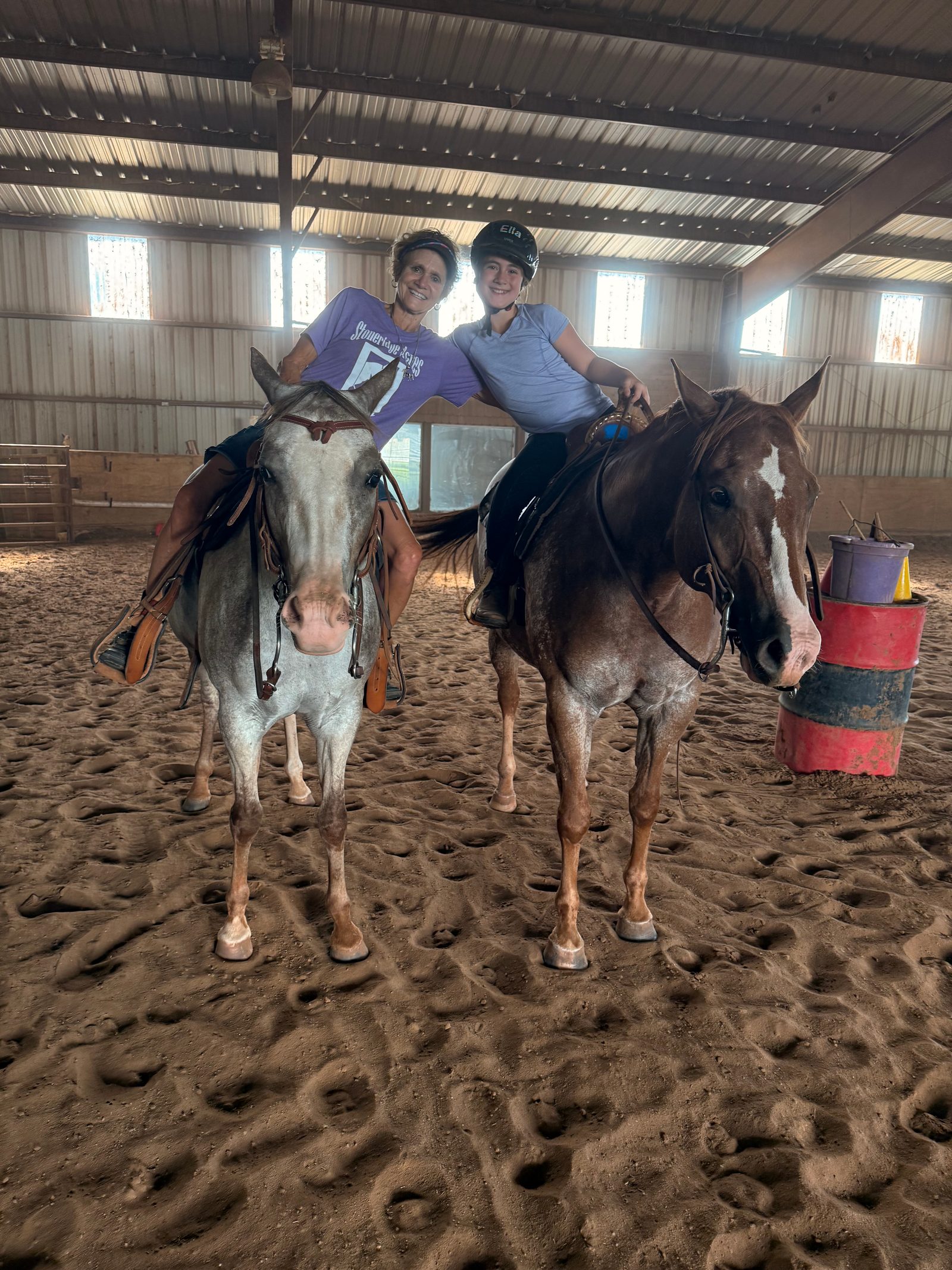 Two smiling riders sit on Appaloosa horses in the indoor arena, one leaning on the other's shoulder.