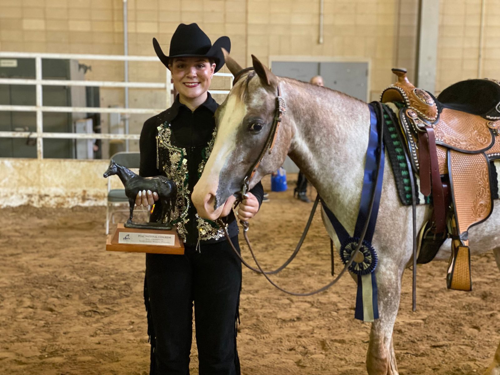 A rider in a black hat stands with her Appaloosa and a championship trophy.