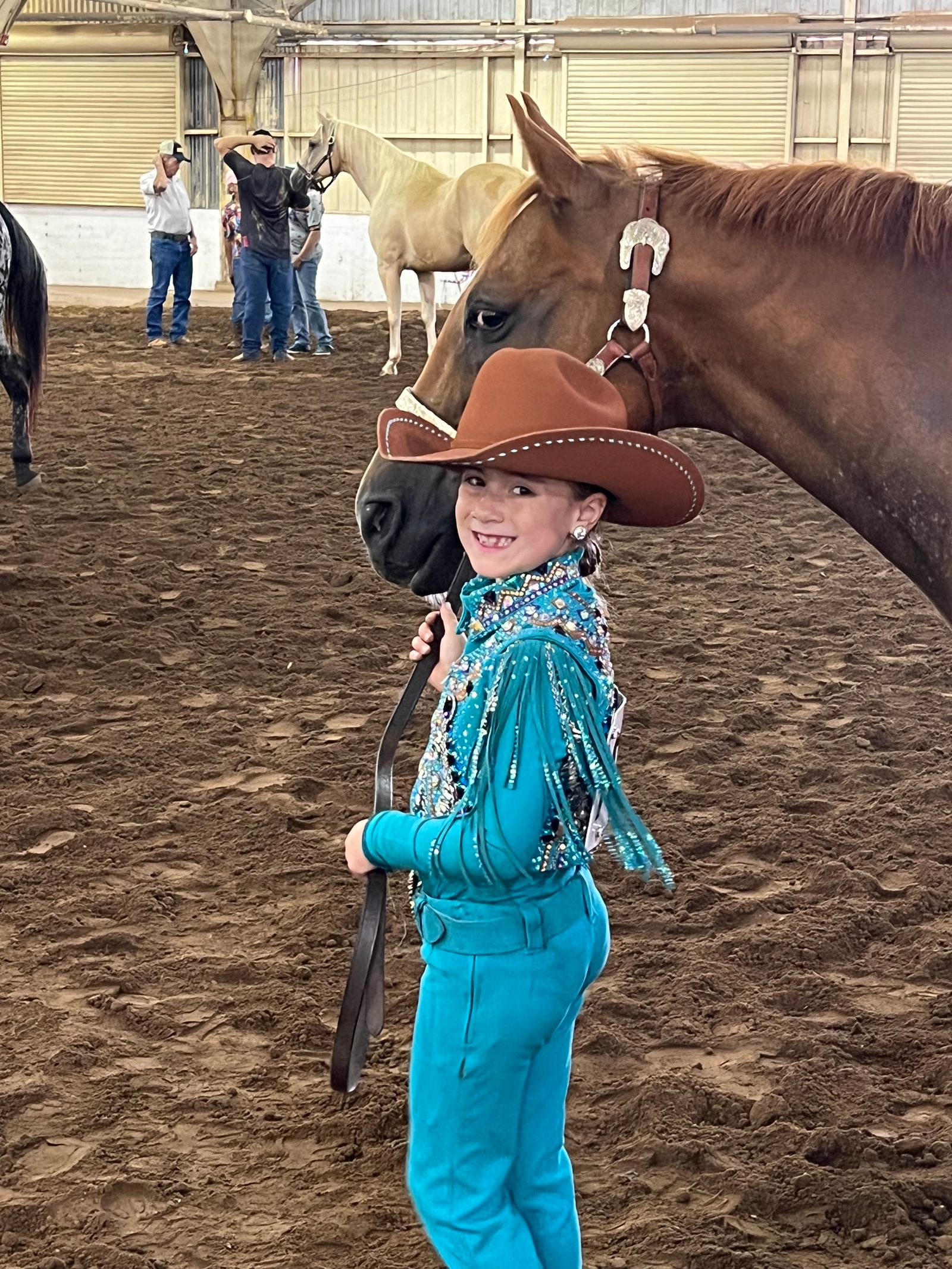 A young rider in a teal show outfit and brown cowboy hat stands with her sorrel horse in the show arena.