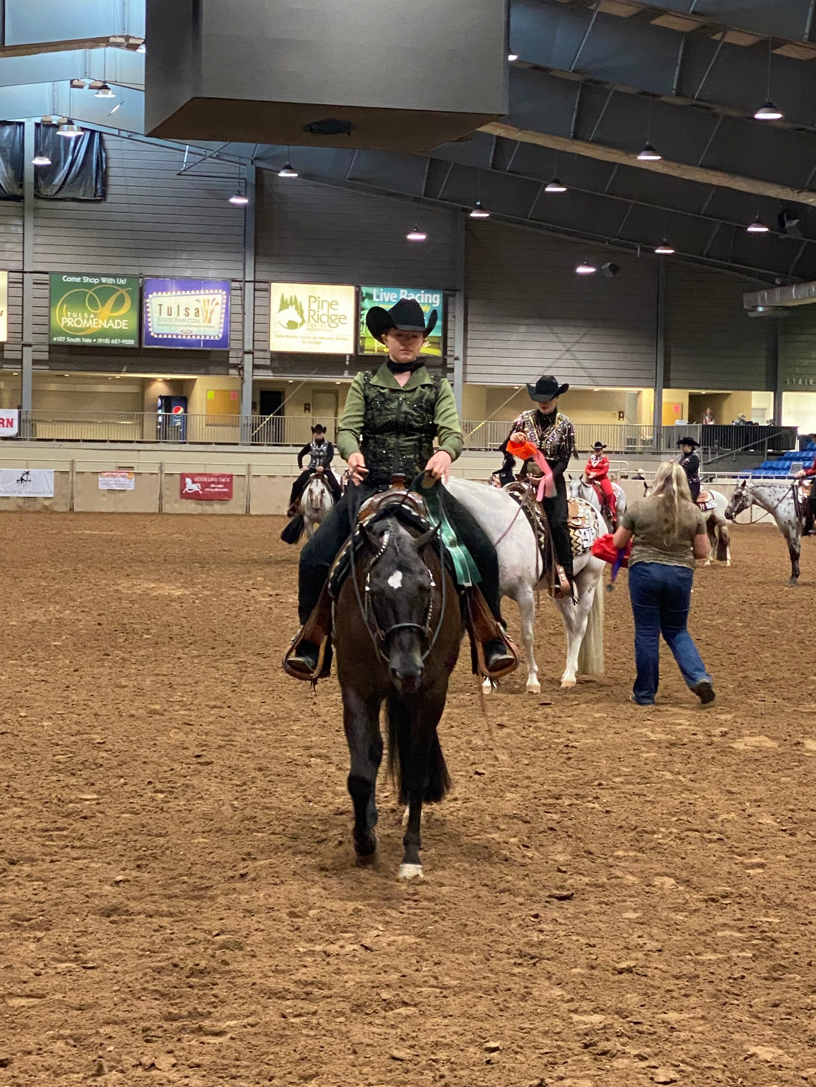A rider in a green western show outfit and black hat rides a dark horse in a Tulsa show arena, with other riders behind.