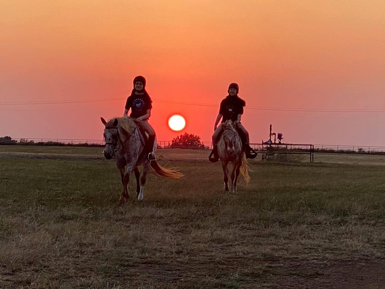 Two riders on Appaloosa horses walk toward the camera across a pasture as the sun sets behind them.