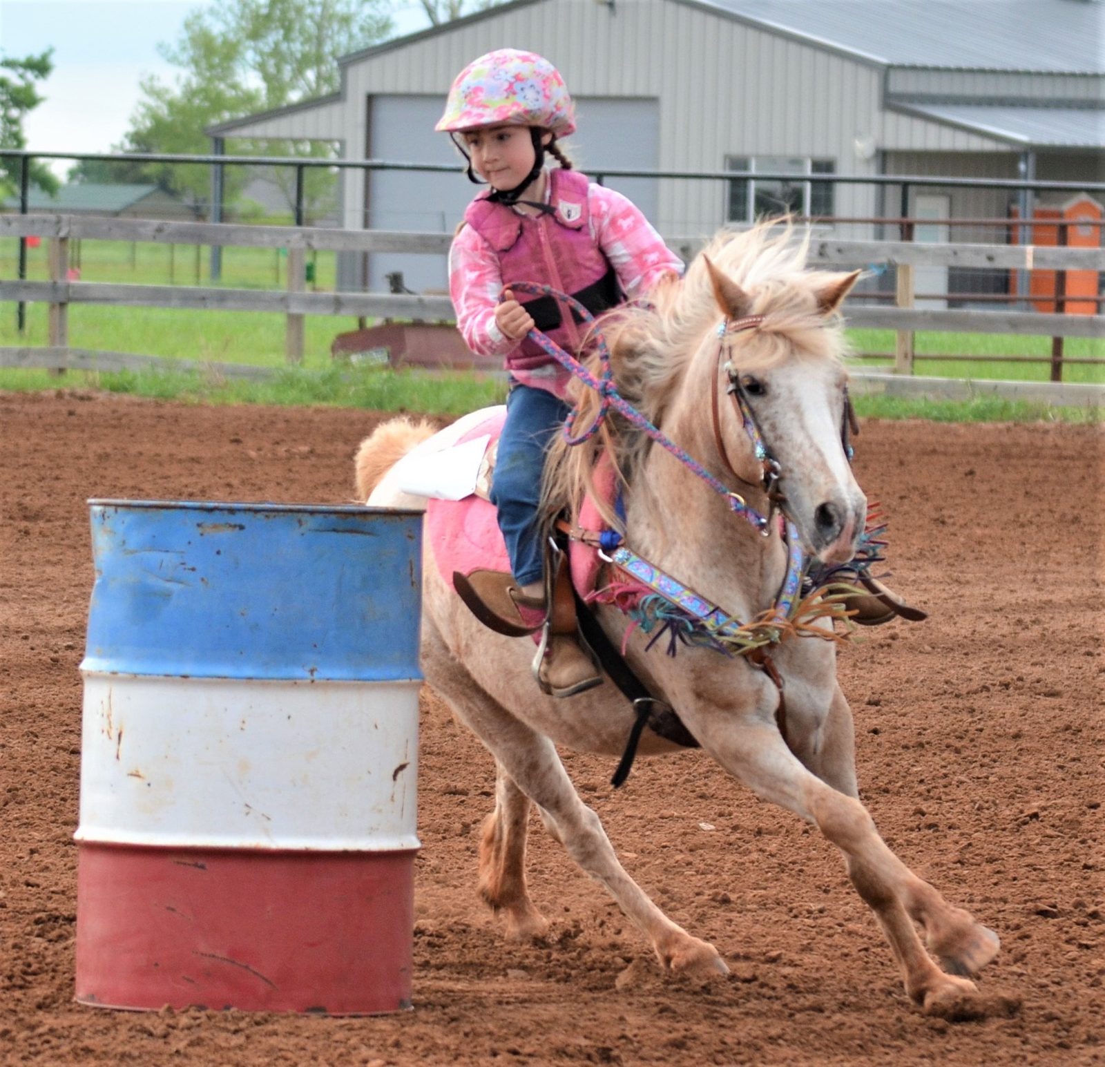 A young rider in a pink helmet rounds a barrel at speed on an Appaloosa.