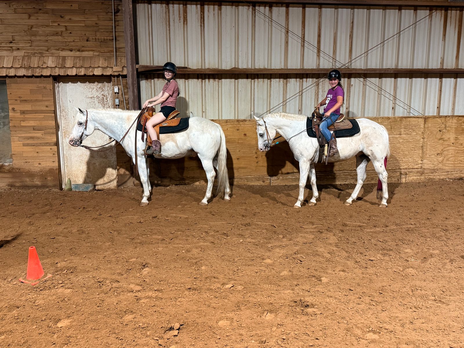Two young riders in helmets sit on grey horses side by side in the indoor arena.