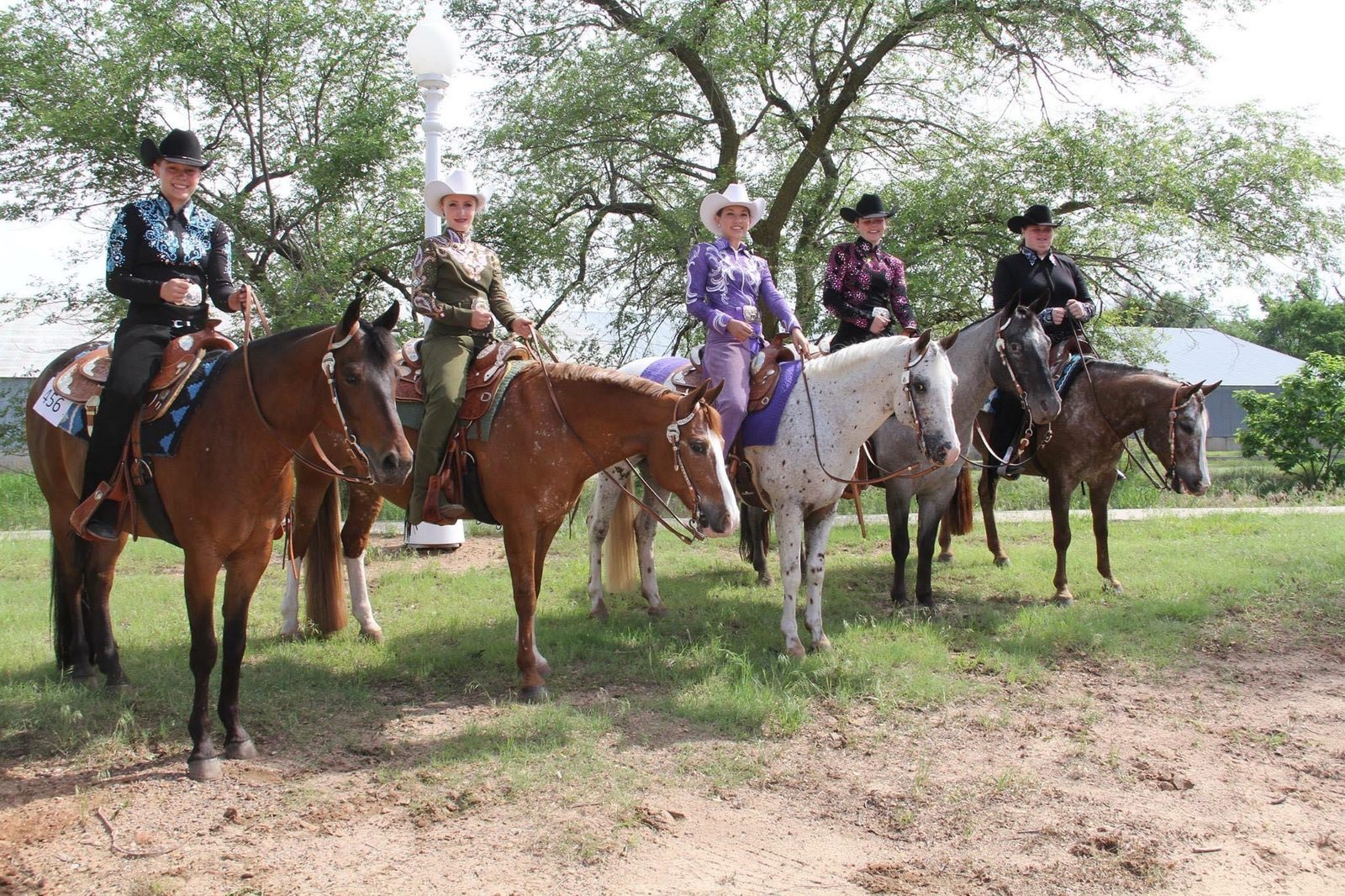 Five riders in western show attire sit on their horses in a line across a green pasture at the barn, with trees behind them.