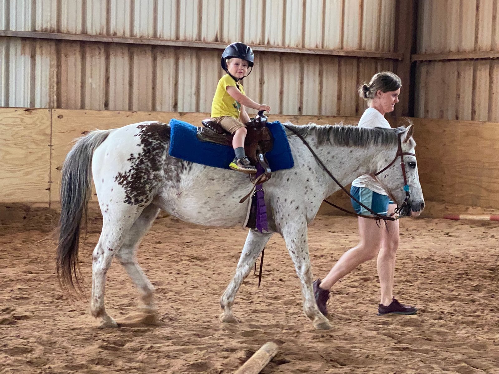 An instructor walks beside a small child riding an Appaloosa in the indoor arena.