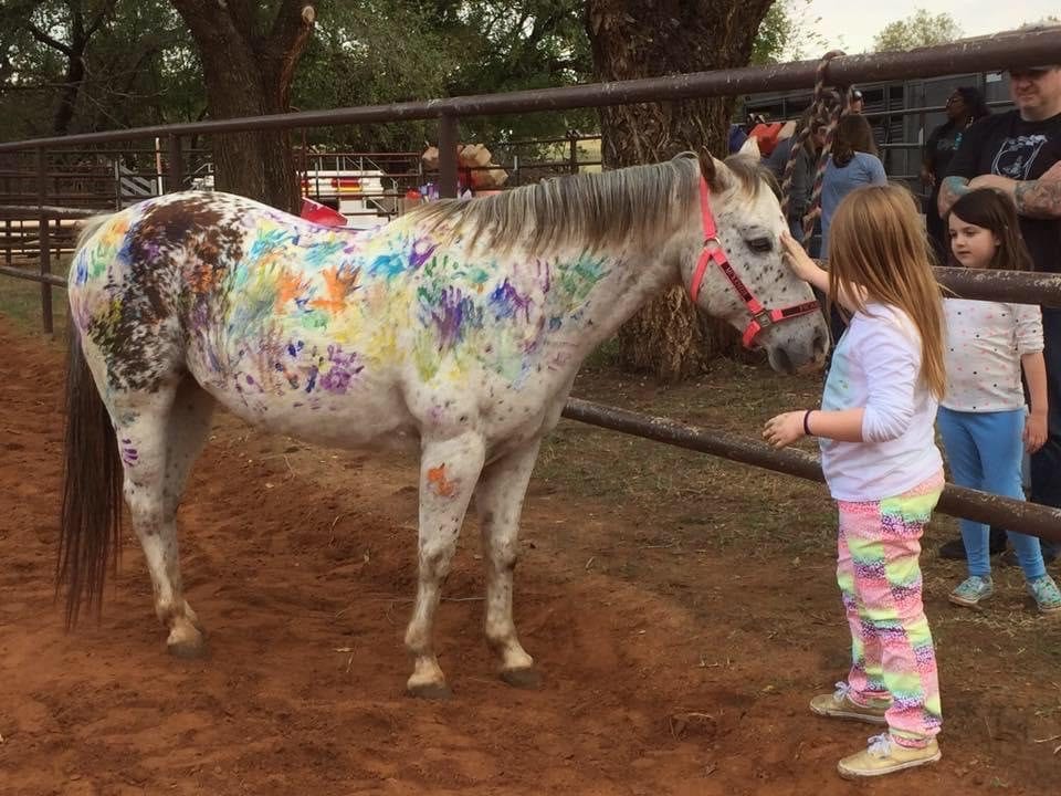 A white pony painted with colorful handprints and designs stands beside children at a birthday party.