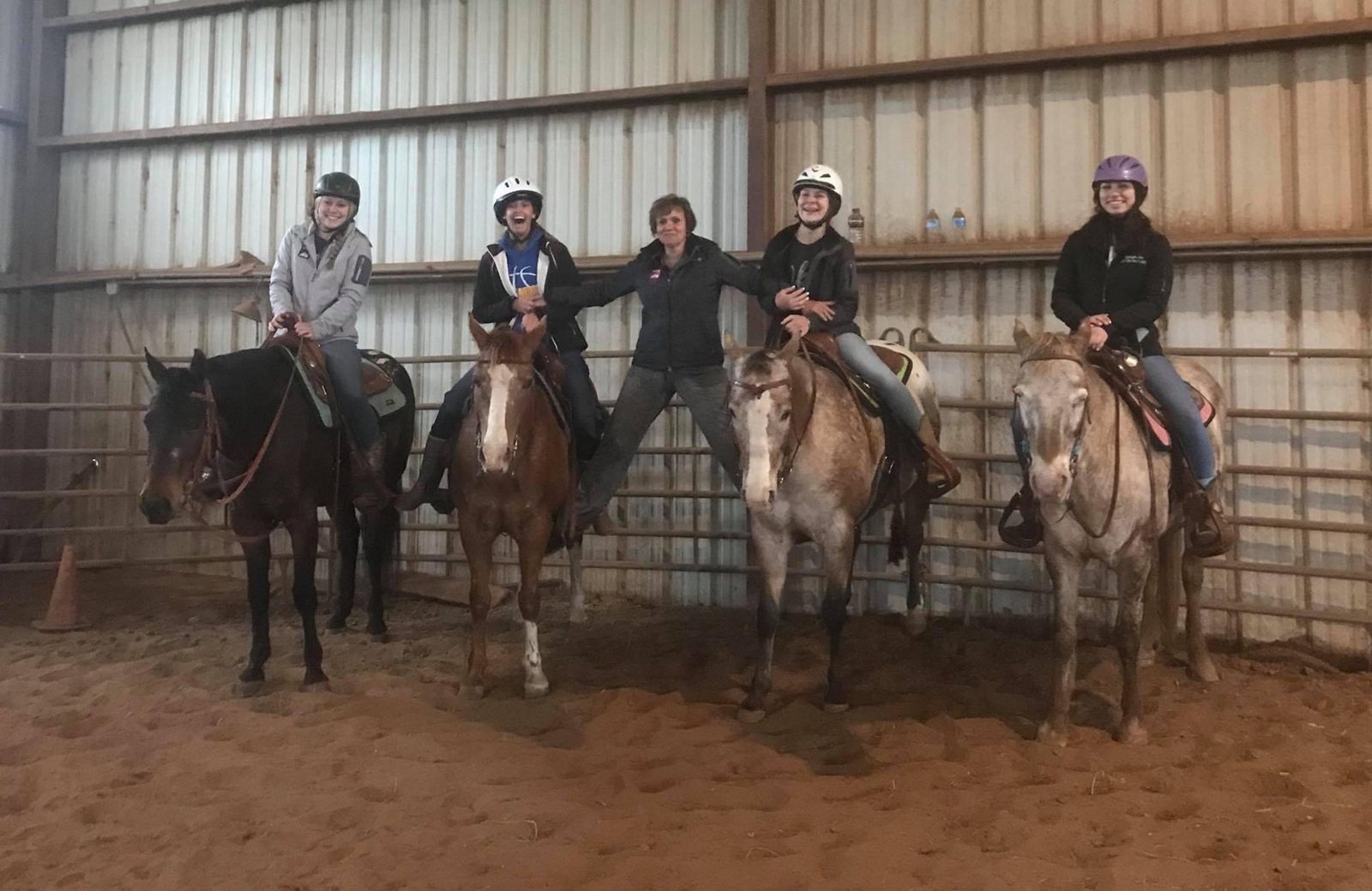 Kathy McKenzie stands in the covered arena between four mounted riders, all laughing.